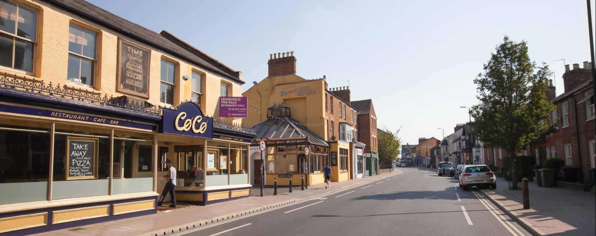 A view of a sunny street with cafes and a tree, this is Cowley Road in Oxford